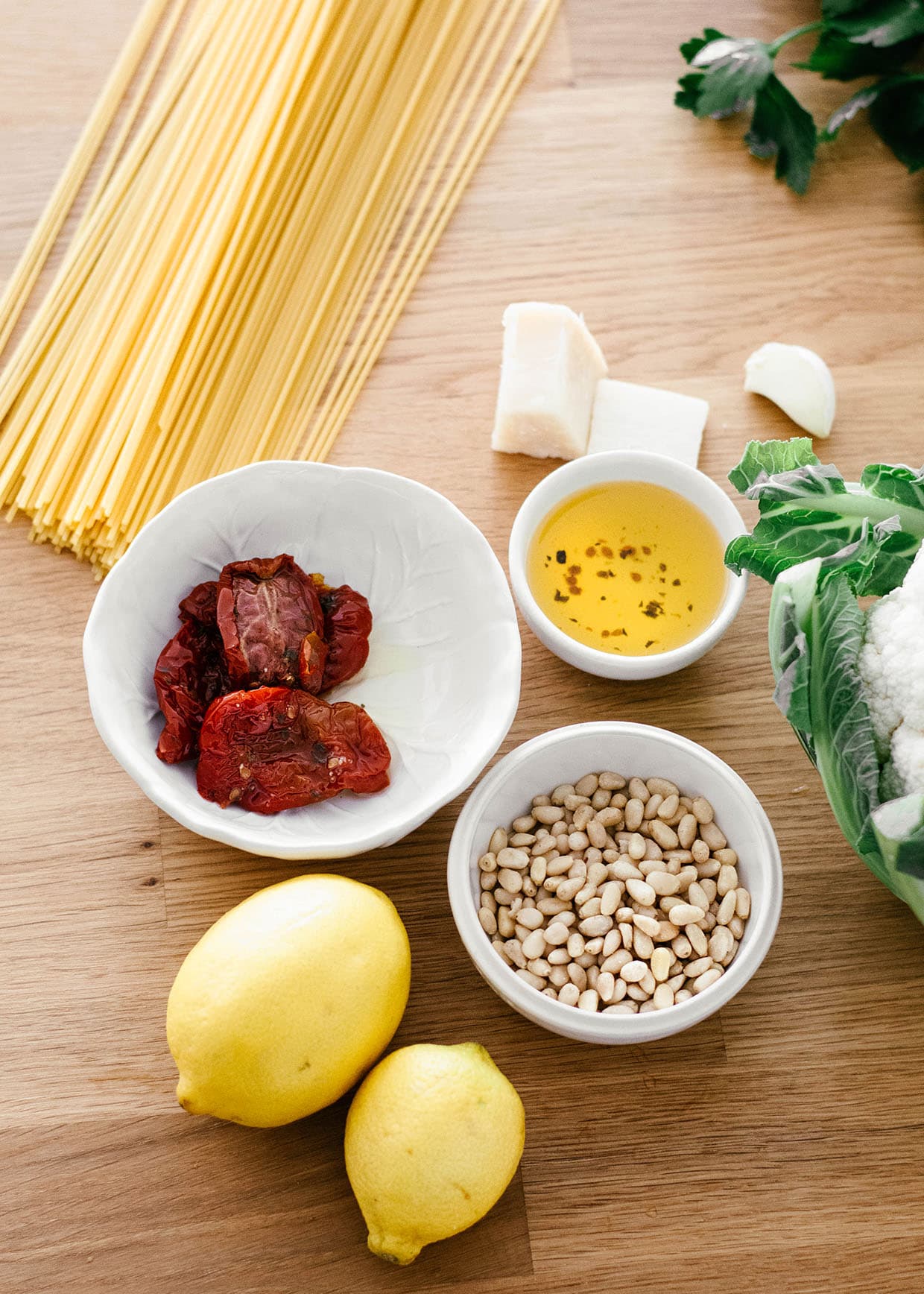 cauliflower pesto ingredients on a table - lemons, pine nuts, sun dried tomatoes, oil, cheese, spaghetti, fresh parsley and cauliflower 