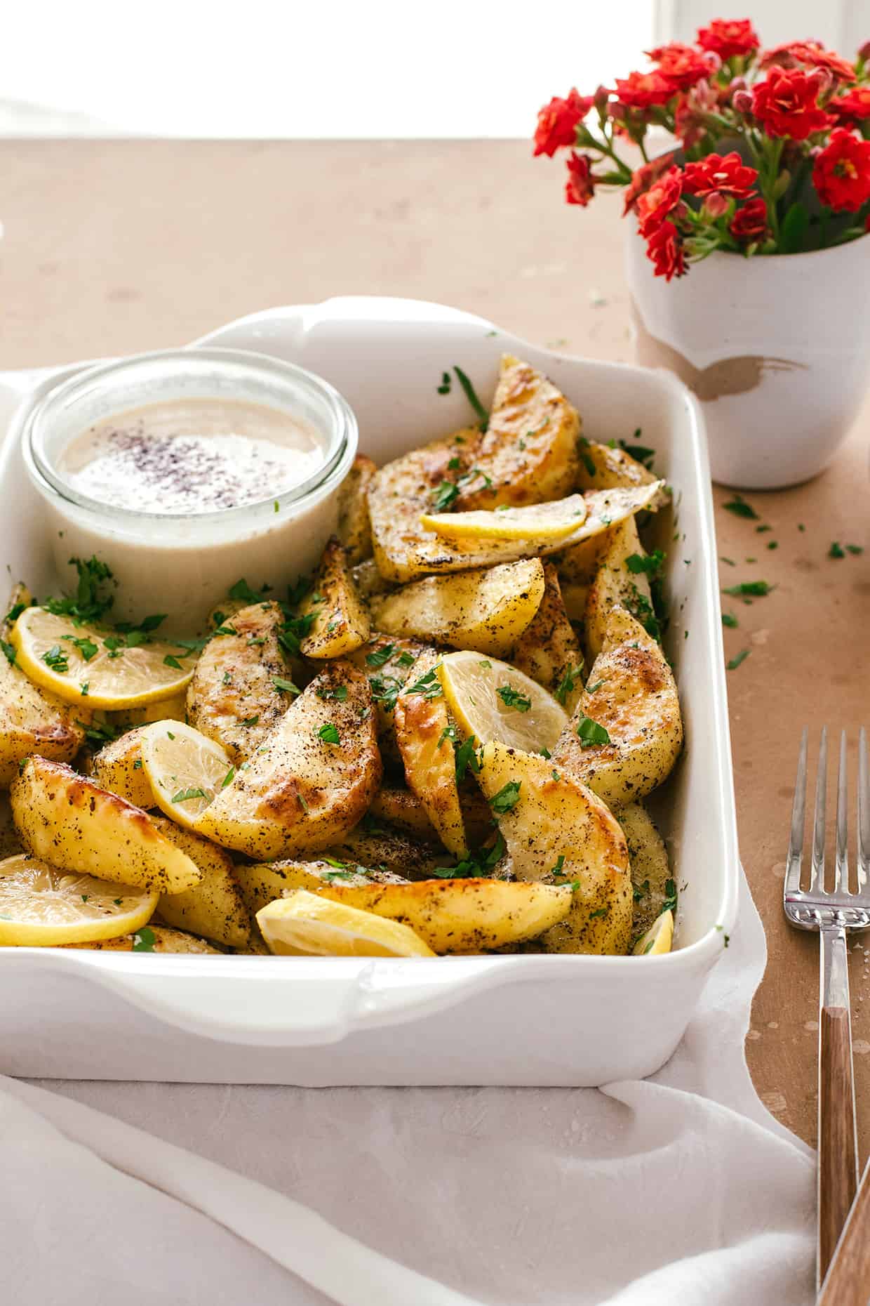 crispy baked potatoes in a baking tray sprinkled with parsley and a glass bowl of tahini sauce