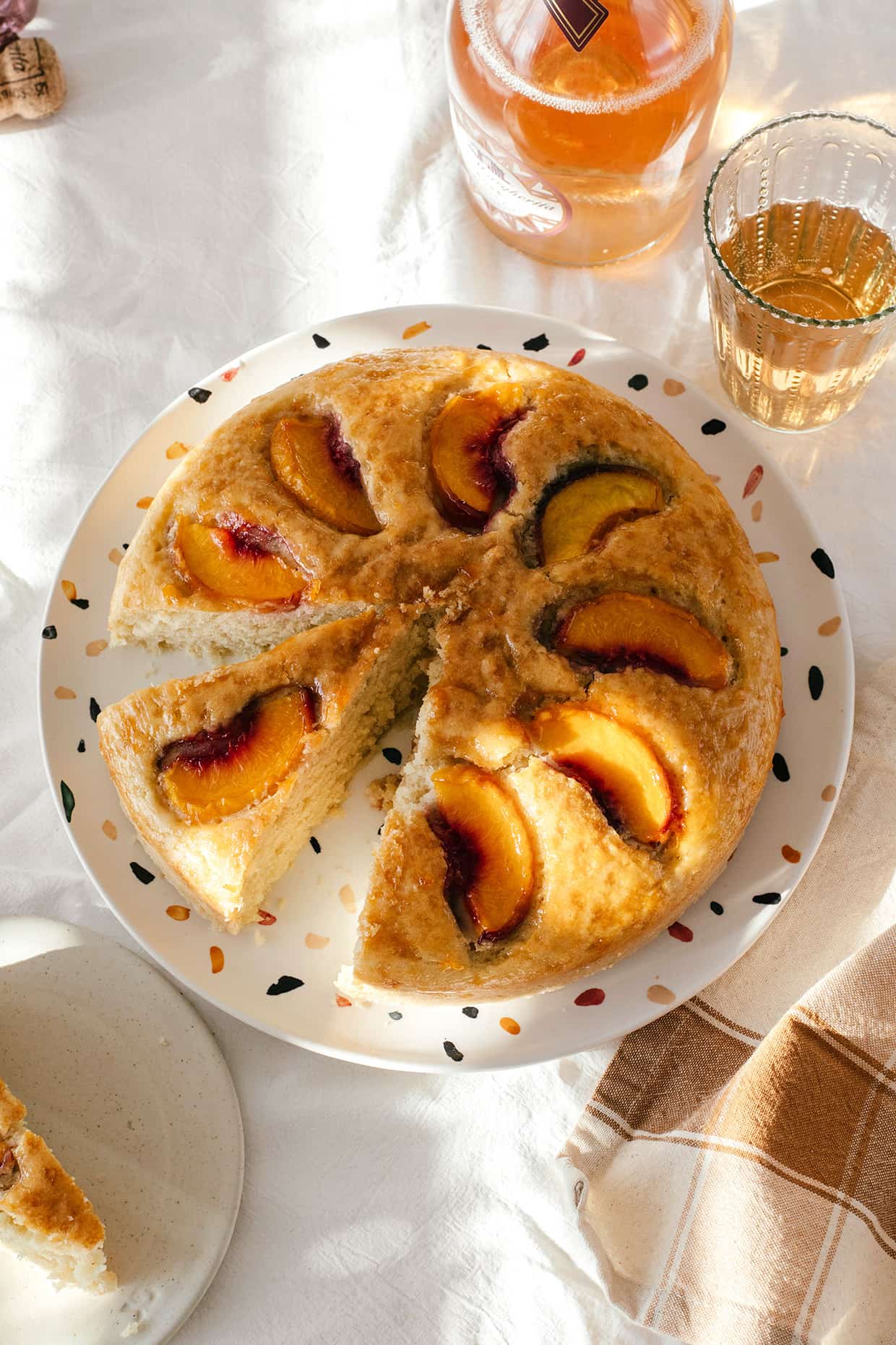 a plate with single layer peach cake, sliced, with a bottle of wine and glasses on the side