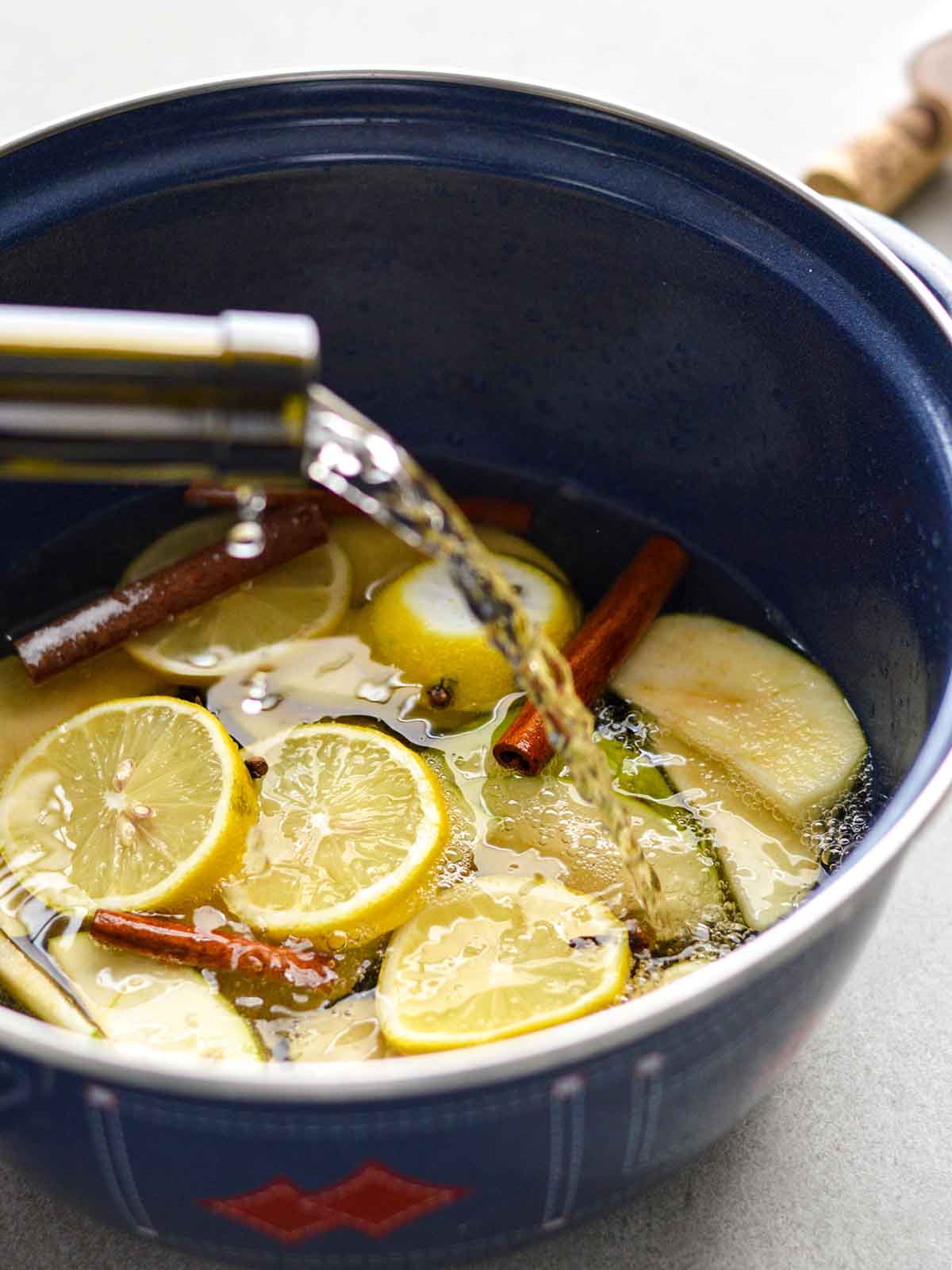 pouring wine into a pot with sliced lemons, apples, cinnamon sticks